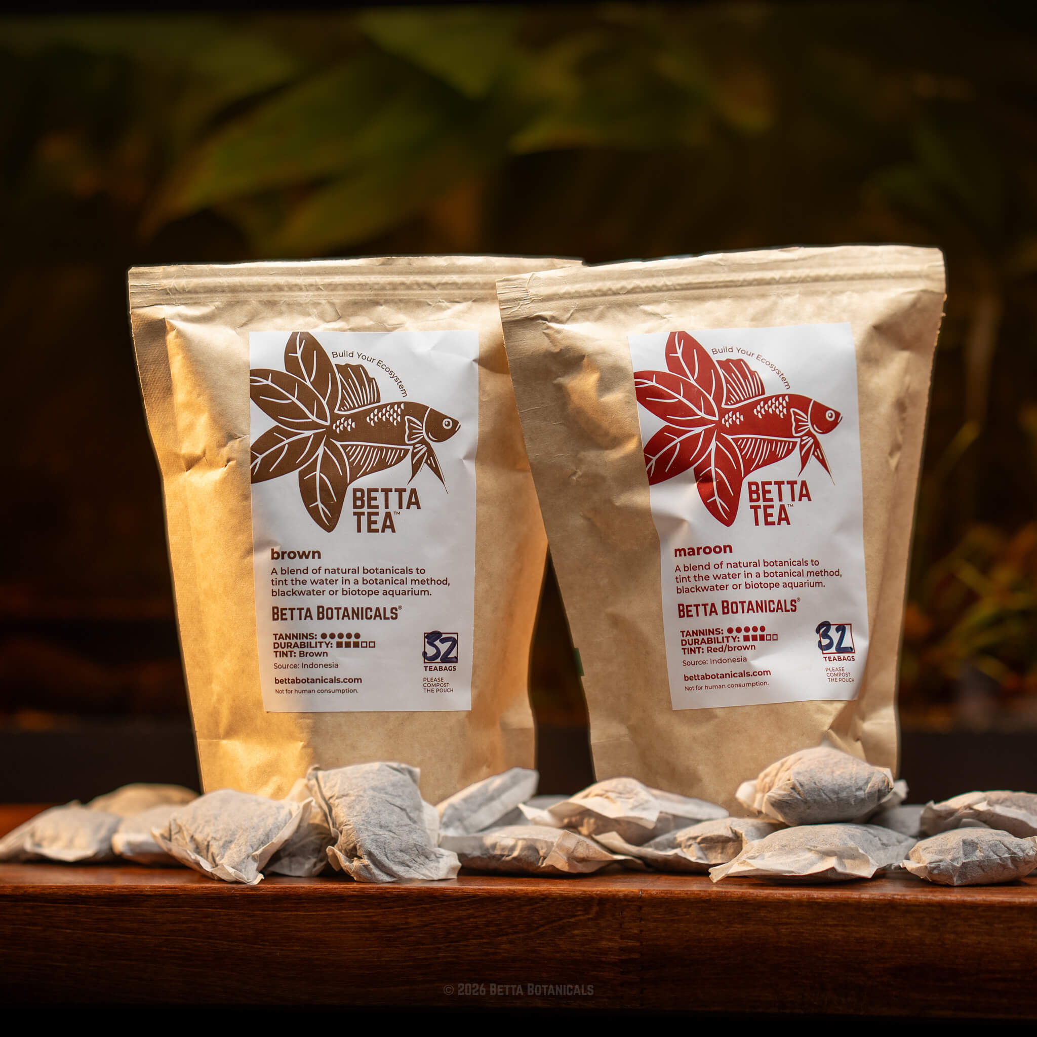 A side by side group photo of brown and maroon betta tea for aquariums in kraft brown compostable pouches and paper tea bags on a wooden desk, photographed by Alex K for Betta Botanicals.