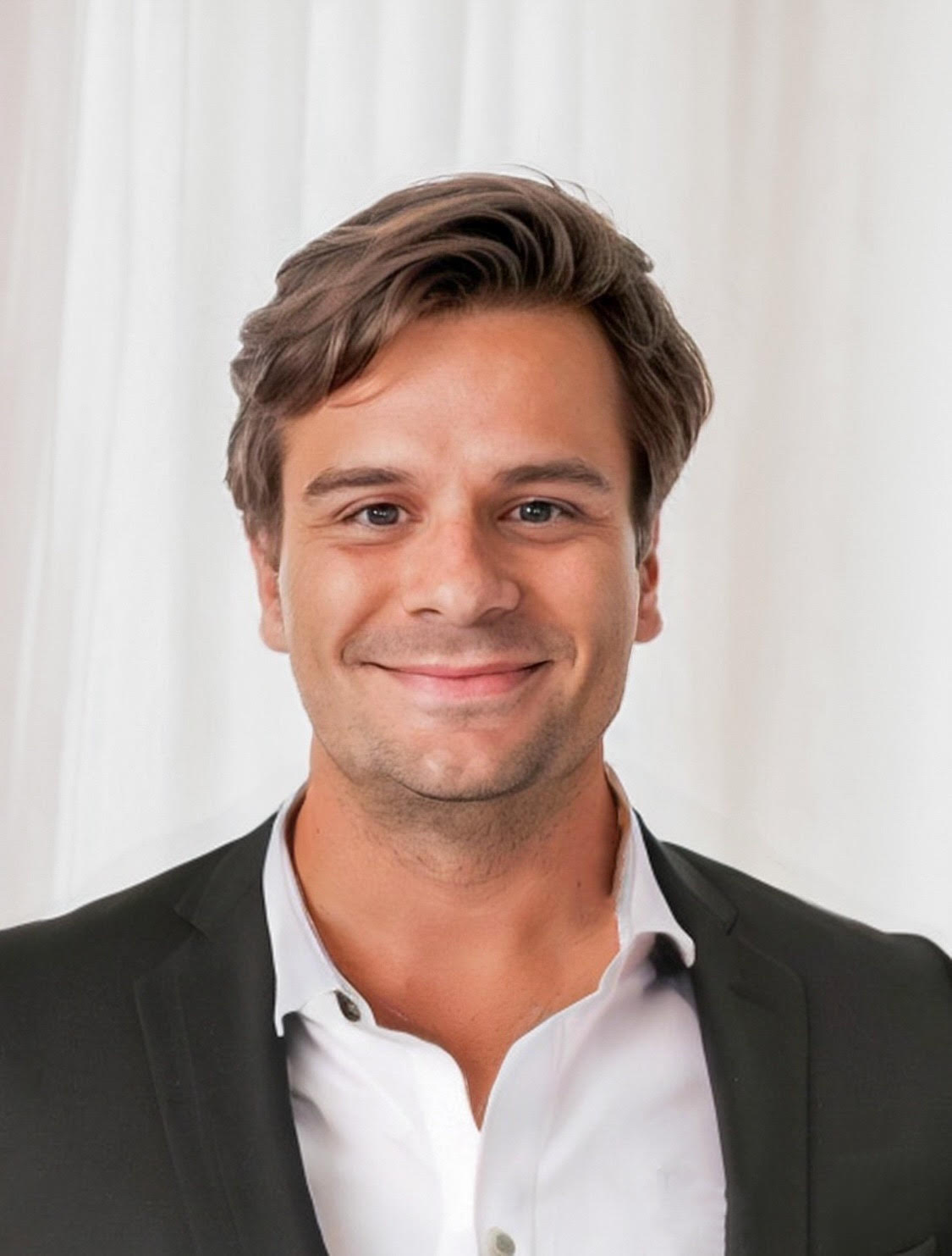A brown haired man with brown eyes in a white button shirt and black suite jacket against a white background.