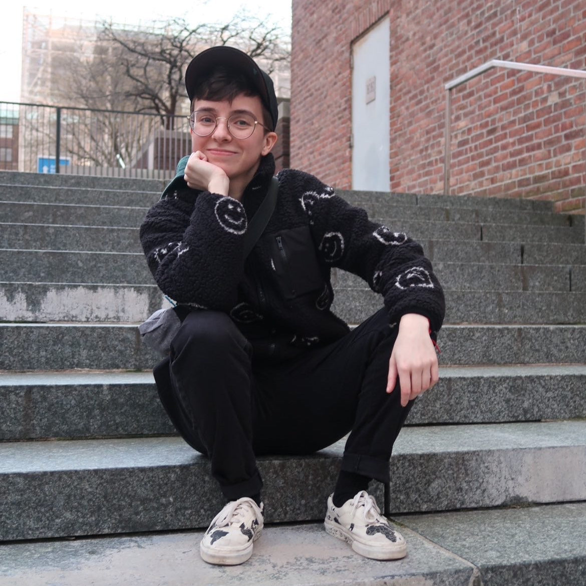 A person sitting on grey stairs in a black hoodie with smiley faces.