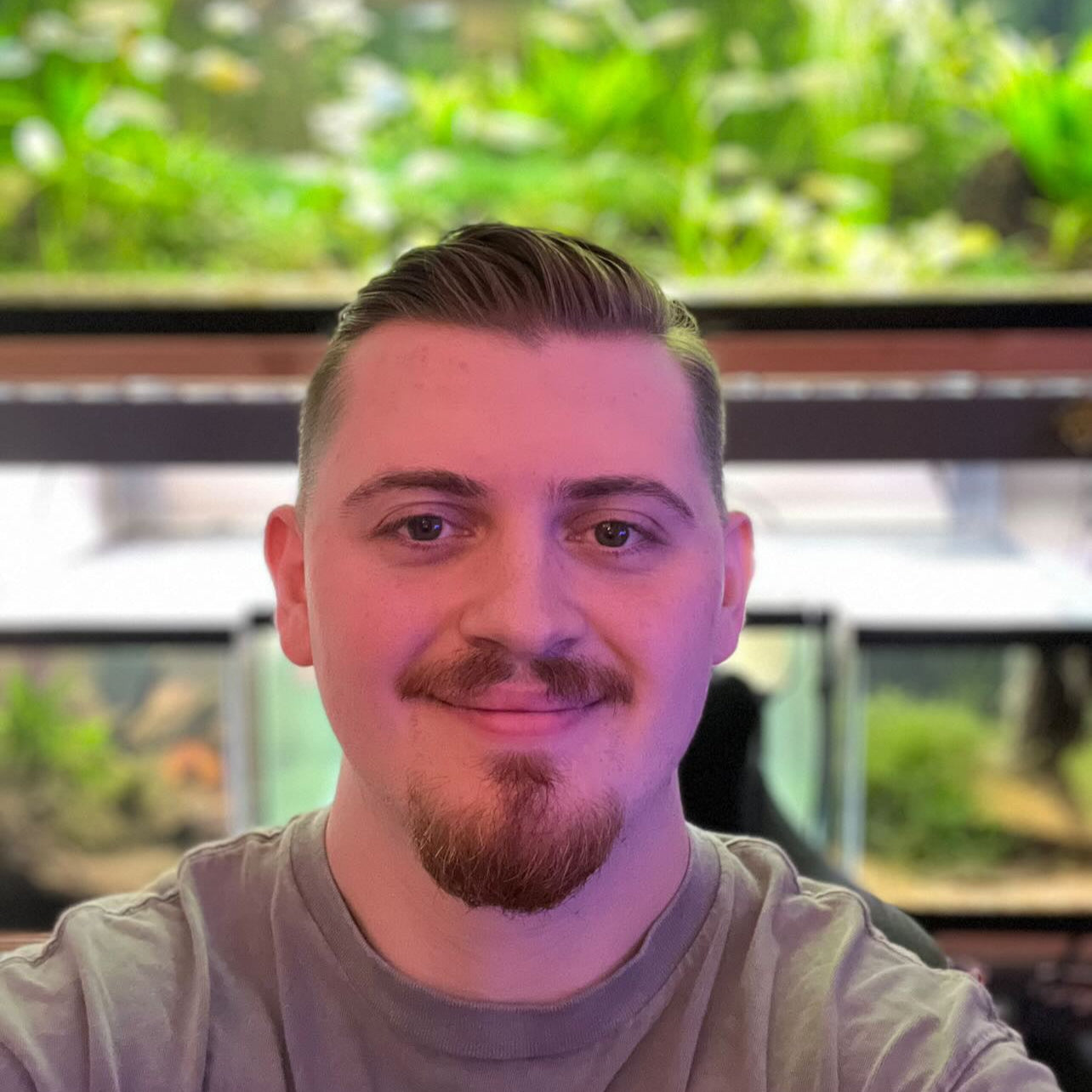 A person with a brushed back hair cut and facial hair in front of five green planted aquariums.